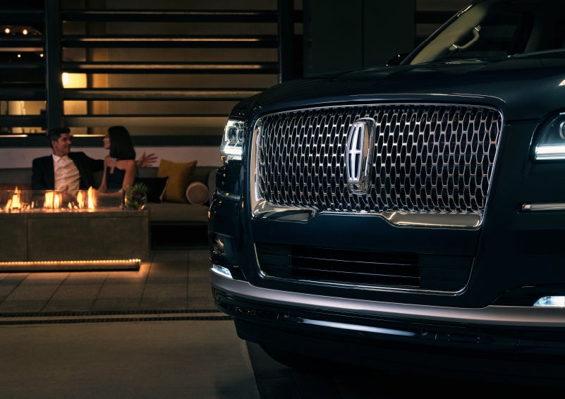 The signature Lincoln grille on the 2022 Lincoln Navigator SUV commands attention as a couple relaxes by a fire beyond | Klaben Lincoln of Warren in Warren OH