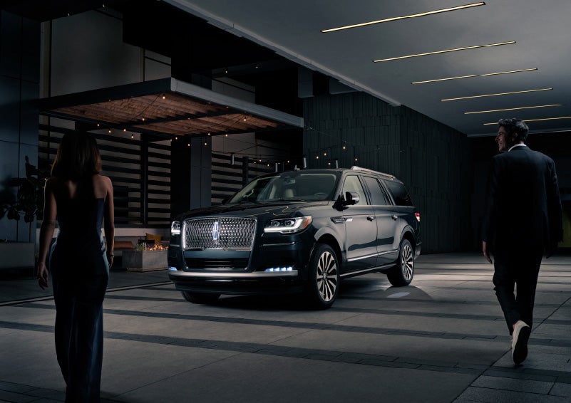 A man and woman in formal wear are approaching a 2022 Lincoln Navigator SUV at night as it lights with the Lincoln Embrace | Klaben Lincoln of Warren in Warren OH