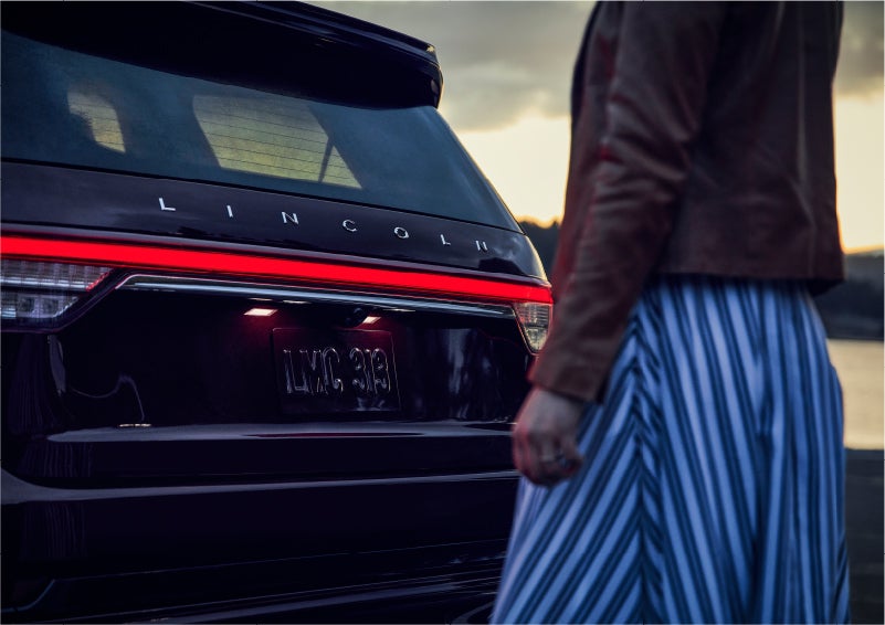 A person is shown near the rear of a 2023 Lincoln Aviator® SUV as the Lincoln Embrace illuminates the rear lights | Klaben Lincoln of Warren in Warren OH