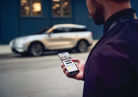 A man is holding his phone looking at the Lincoln Way™ App and we see his 2022 Lincoln Corsair SUV in the background against a blue brick wall | Klaben Lincoln of Warren in Warren OH