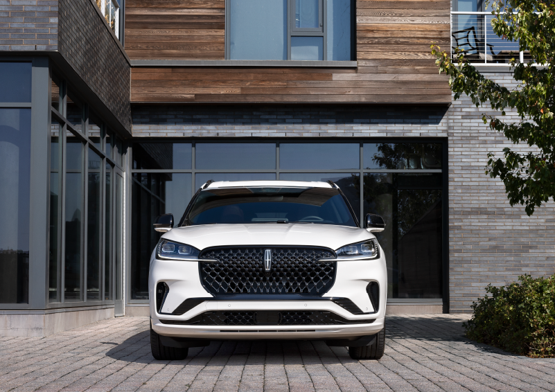 A white 2025 Lincoln Aviator® SUV is shown parked outside a modern building with gray brick and wood accents. | Klaben Lincoln of Warren in Warren OH
