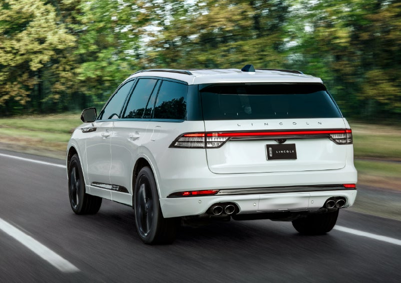A white 2026 Lincoln Aviator® SUV is being driven away down a road lined with colorful trees. | Klaben Lincoln of Warren in Warren OH